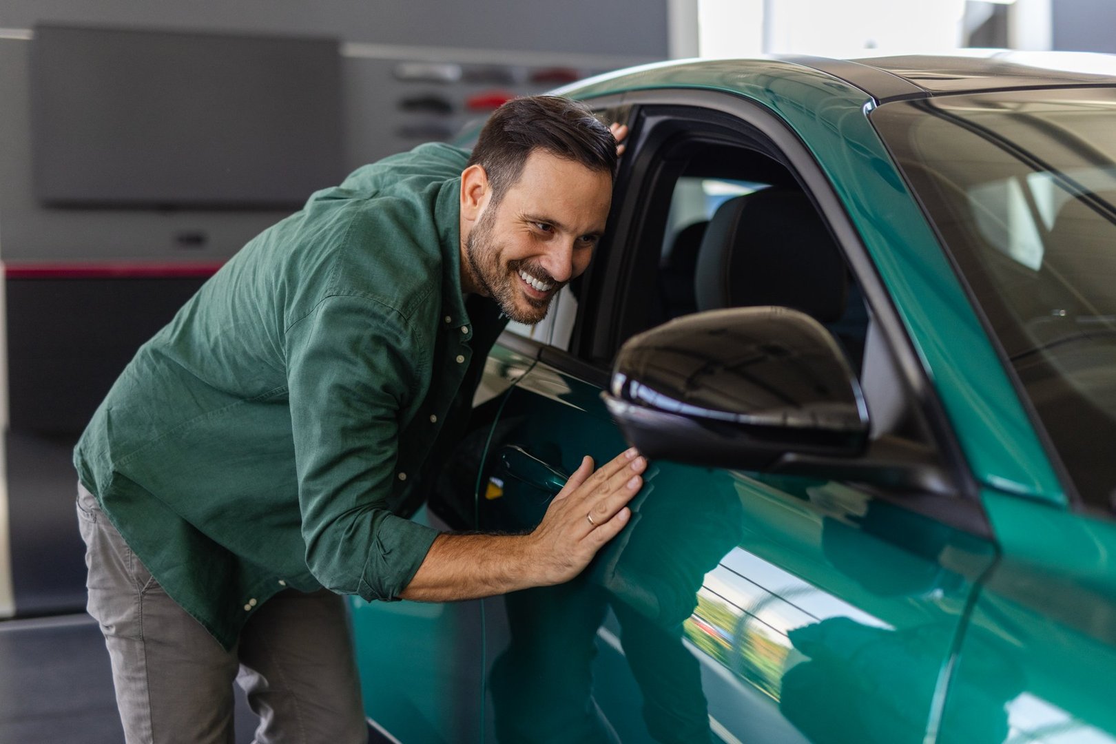 Happy young man hugging his new car in showroom. Satisfied guy with closed eyes embracing the hood of the automobile. Dreaming man lying on car bonnet hugging it.