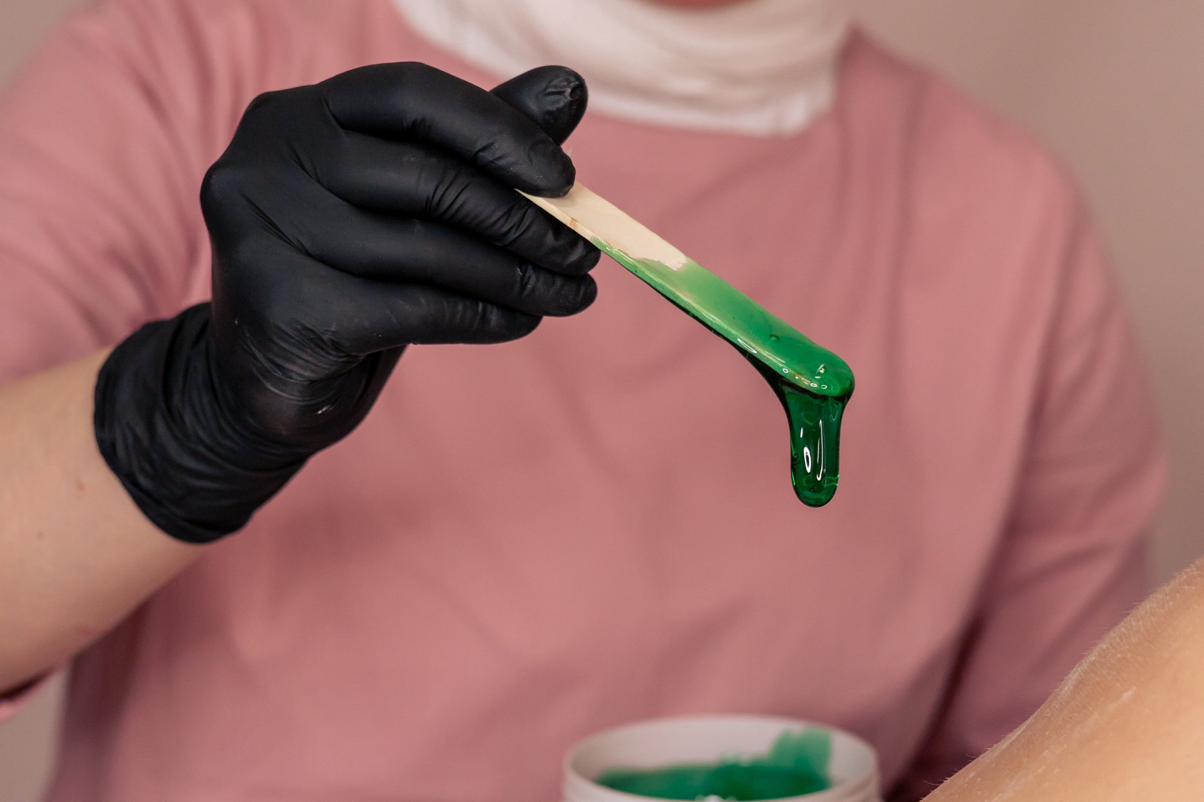 A beautician in gloves carefully applies warm green wax to a clients skin in a beauty salon. The setting reveals a clean and organized workspace, emphasizing the professional atmosphere of the waxing process. The therapist holds a wooden stick, ensuring an even spread of the wax in preparation for hair removal.