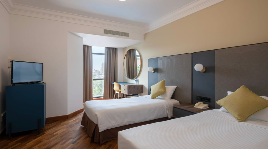 Modern hotel room with two single beds, a TV, desk, and city view from a large window.