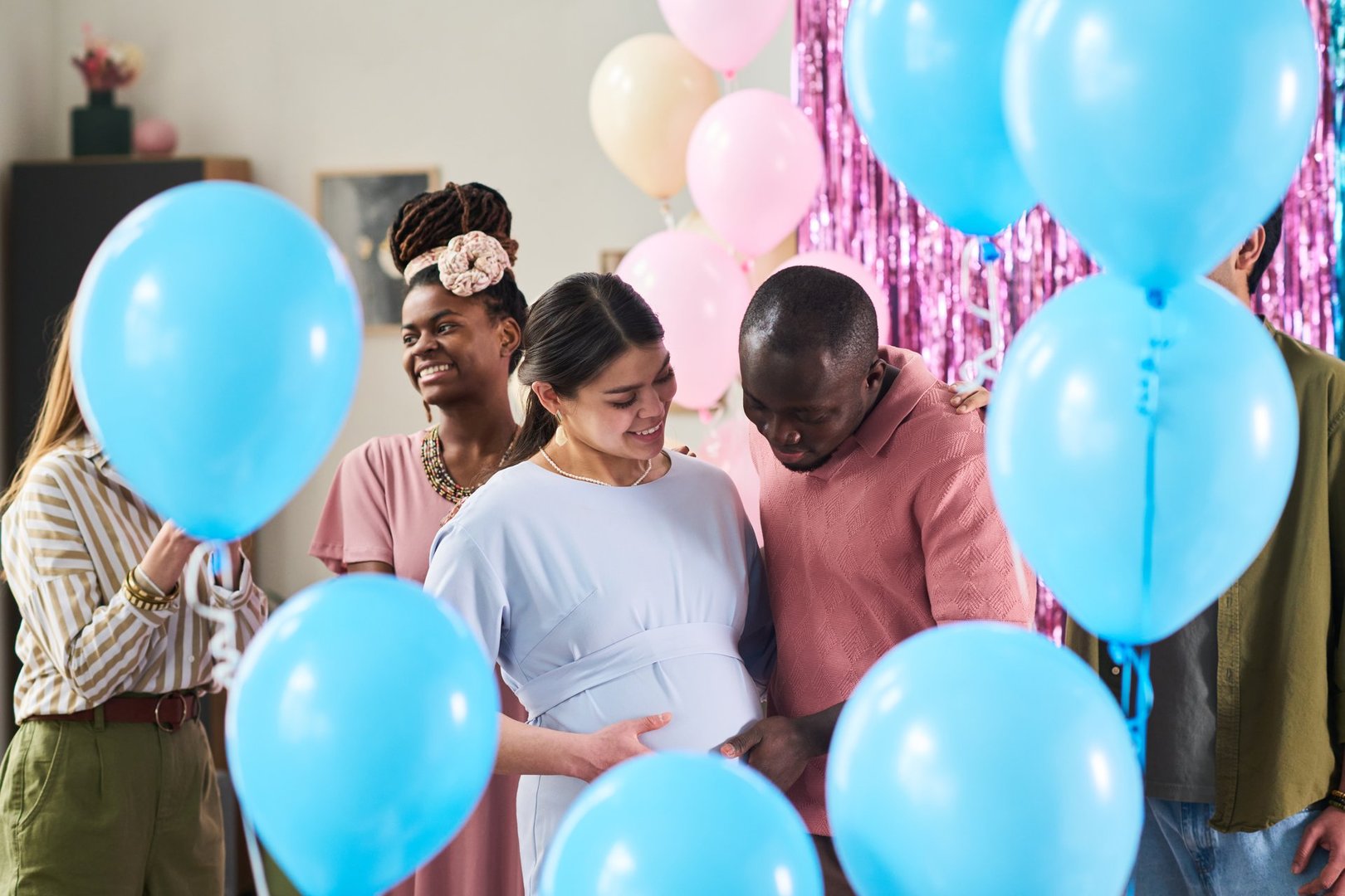 Young adult Caucasian woman with pregnant belly standing beside Black man gently touching her stomach, surrounded by multiethnic friends