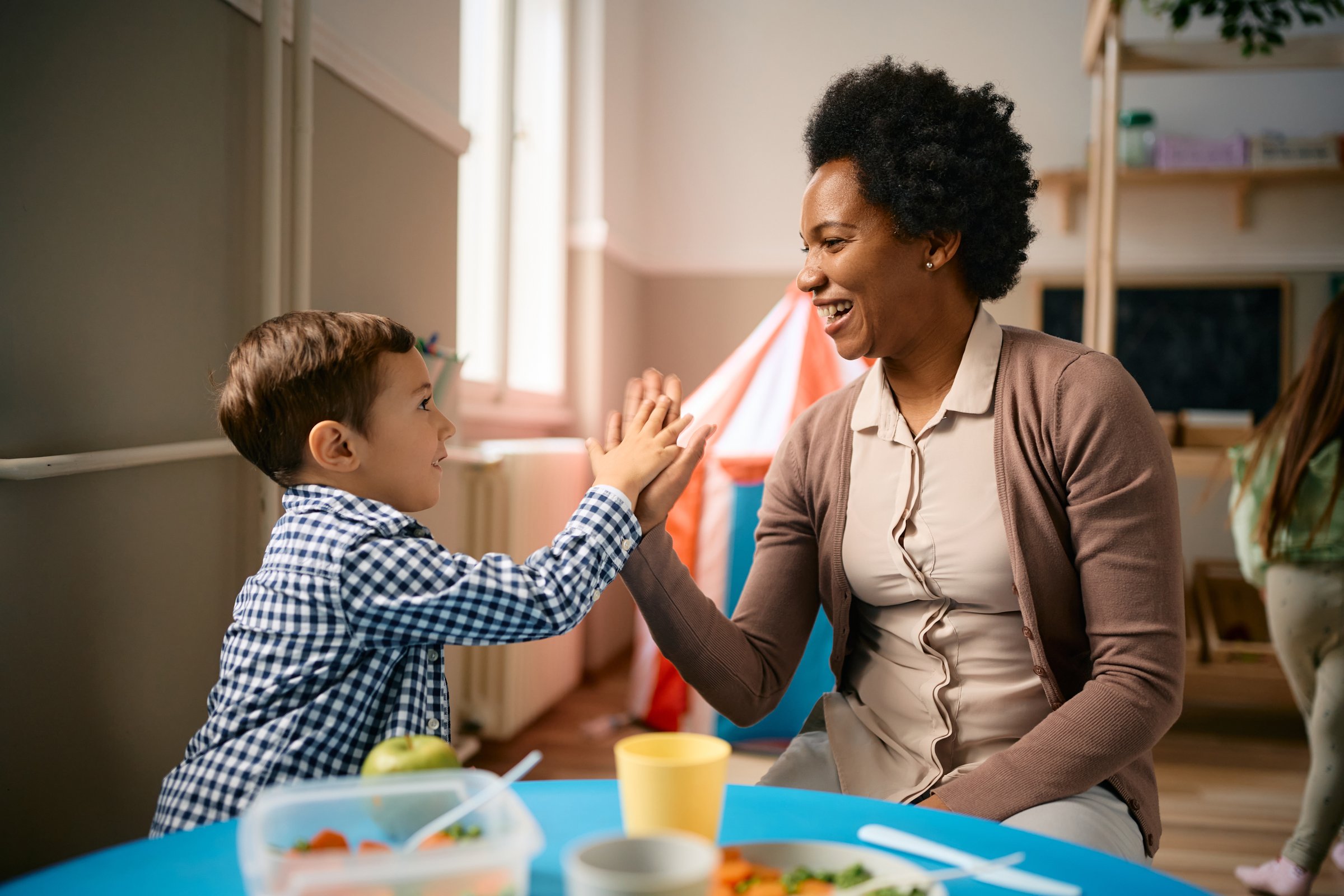 Happy little boy giving high-five to his African American teacher after eating lunch at kindergarten.