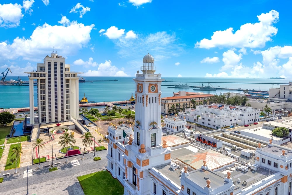 Aerial view of a coastal city with a prominent white clock tower and modern buildings, overlooking a port and the sea.