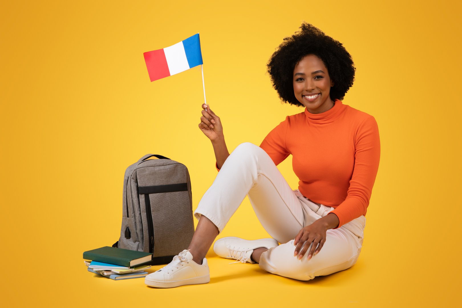 Smiling millennial African American woman with curly hair sitting cross-legged on the floor, holding a French flag, with a backpack and books nearby, against a yellow background