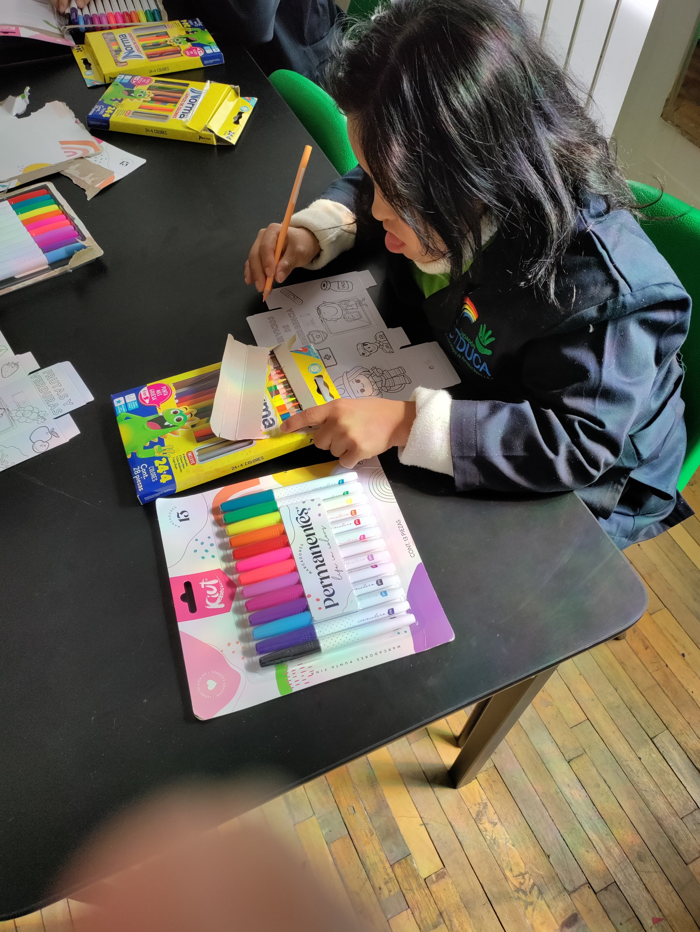 A person coloring on a worksheet at a table with colored pens and markers nearby, seated on a green chair.