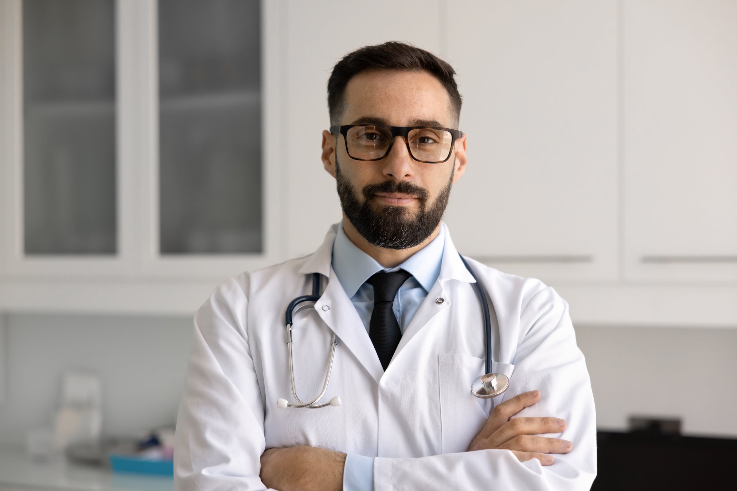 Positive handsome Hispanic medical practitioner man in glasses and lab coat standing in hospital office with arms folded, looking at camera, posing fo doctor professional head shot portrait