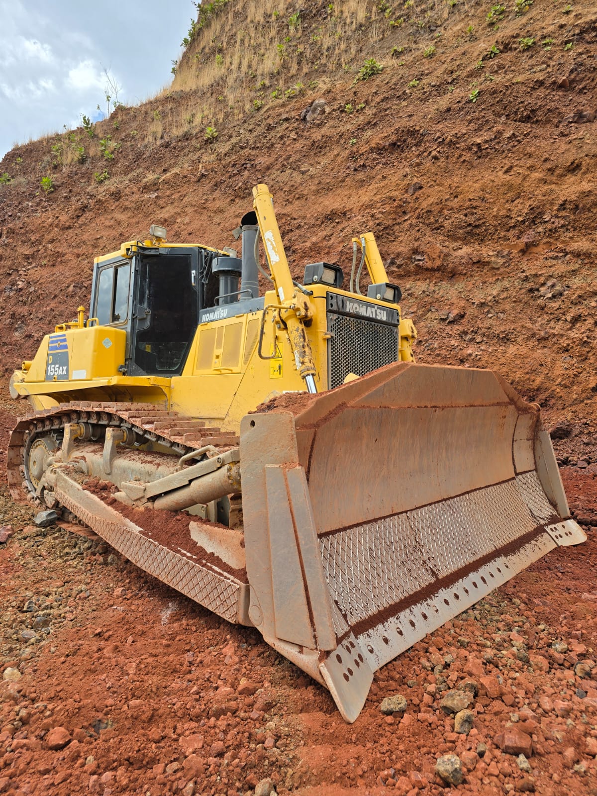 Yellow bulldozer on a red dirt hill, parked with a visible blade, against a backdrop of soil and rock worksite.