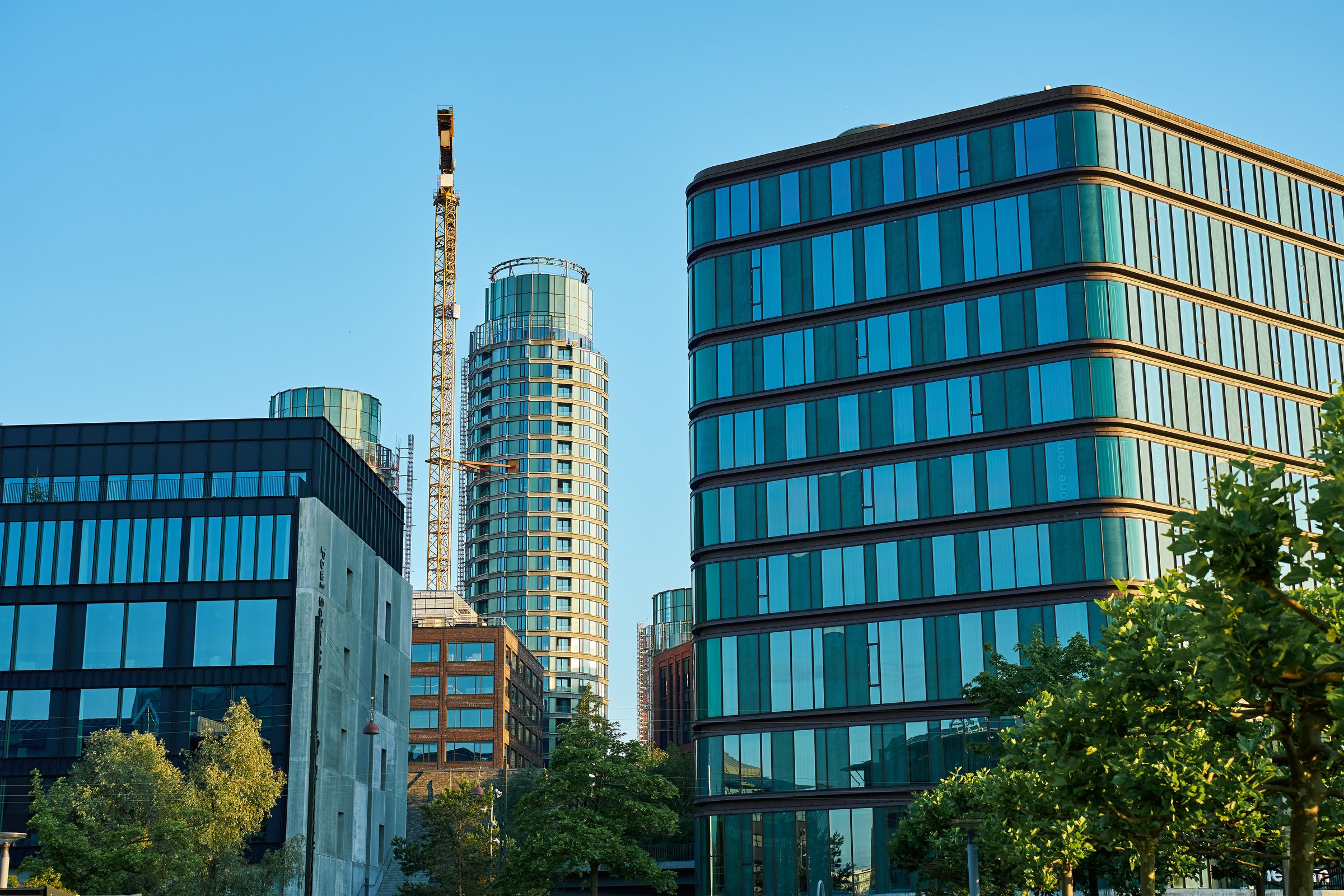 Modern glass buildings with a construction crane surrounded by lush green trees. Sustainable urban development combining contemporary architecture and environmental awareness