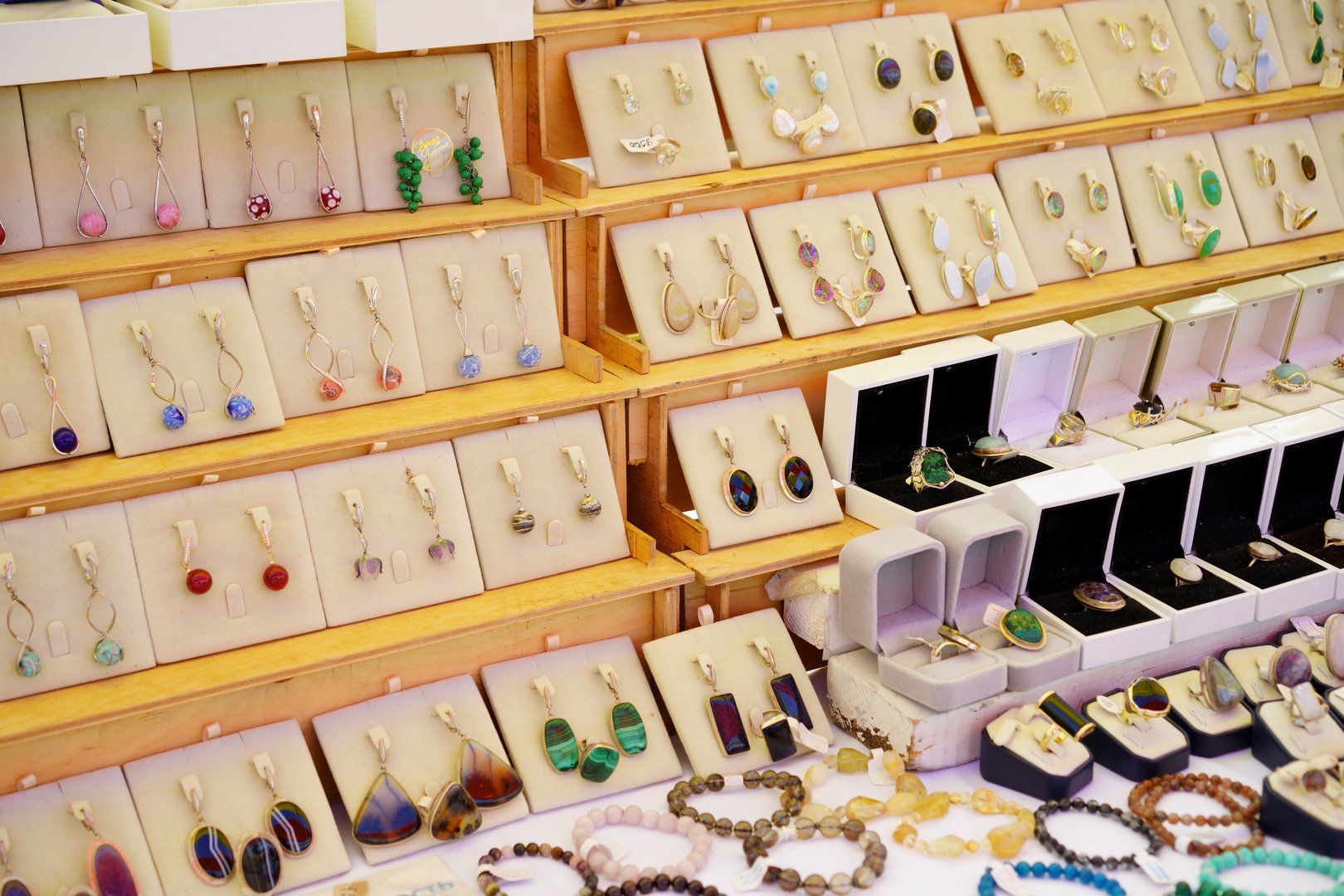 Colorful handmade jewelry is carefully arranged on display stands at a bustling market. Shoppers enjoy browsing the unique earrings, rings, and bracelets on a warm day.