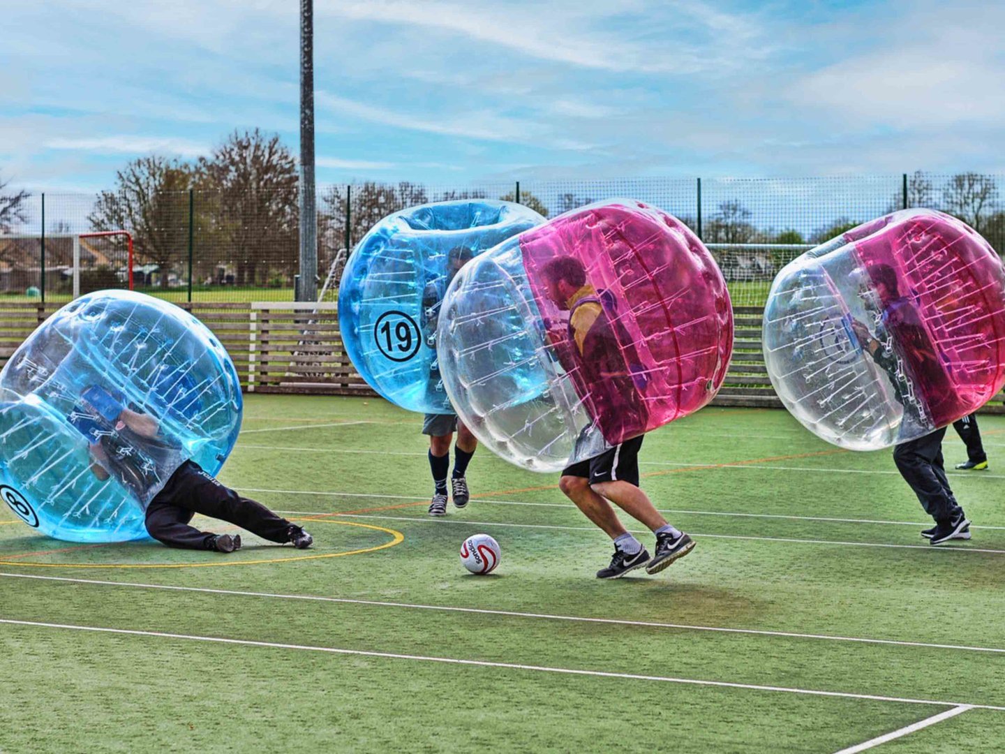 People playing bubble soccer on an outdoor field, wearing inflatable balls for protection, with a soccer ball in play.