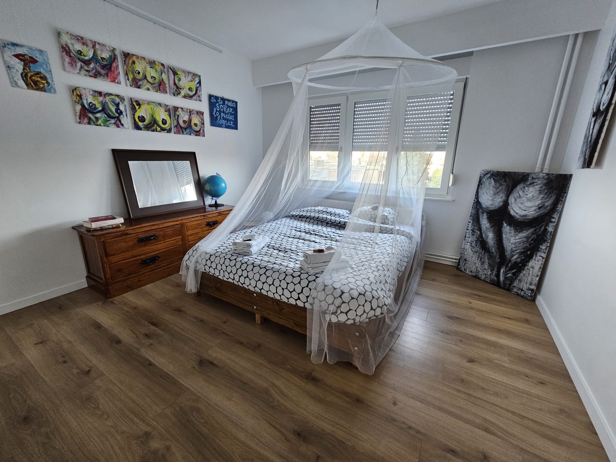 A bedroom with a bed under a canopy, wooden floor, dresser with mirror, globe, abstract paintings, and large windows.