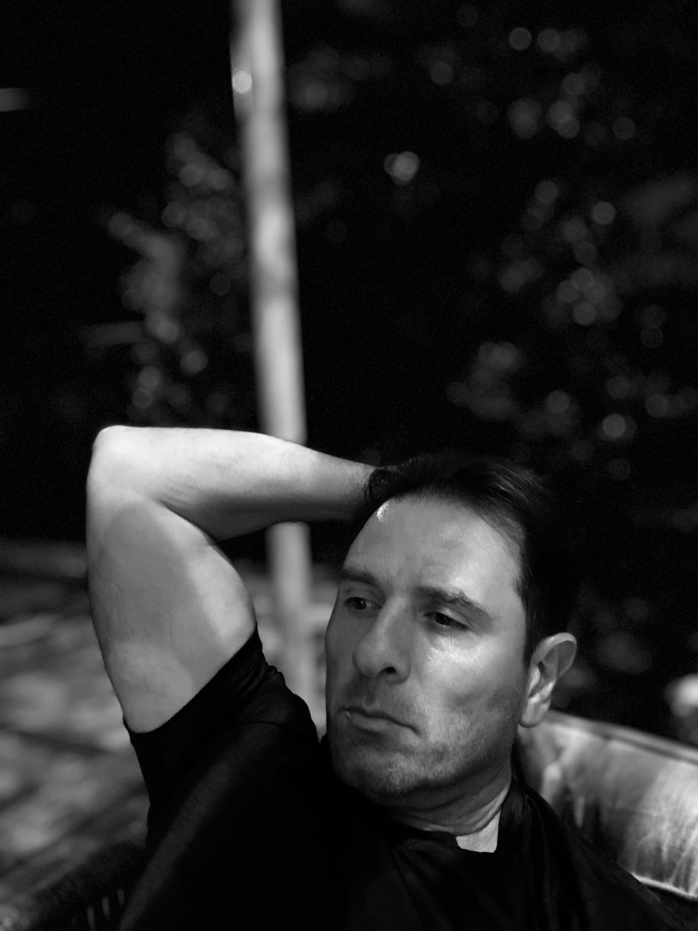 Black and white image of a person sitting outdoors at night with one arm resting behind their head, looking contemplative.