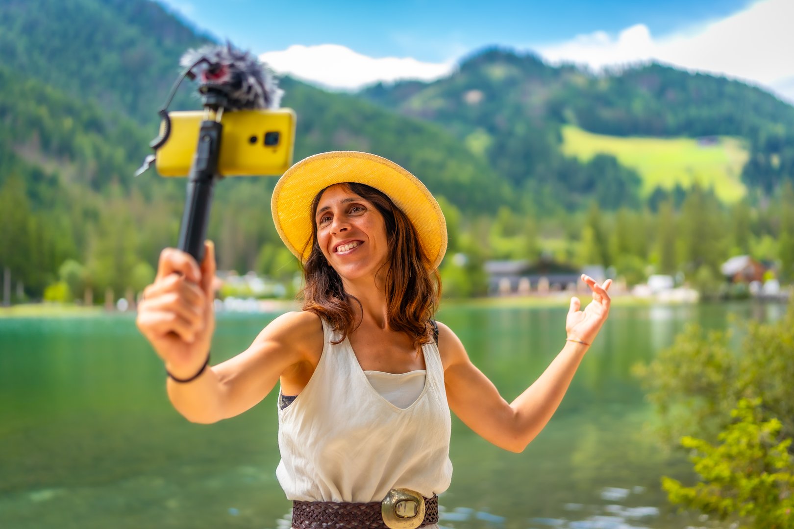 Smiling woman wearing straw hat vlogging with smartphone and external microphone at lake dobbiaco in the italian dolomites during summer holidays