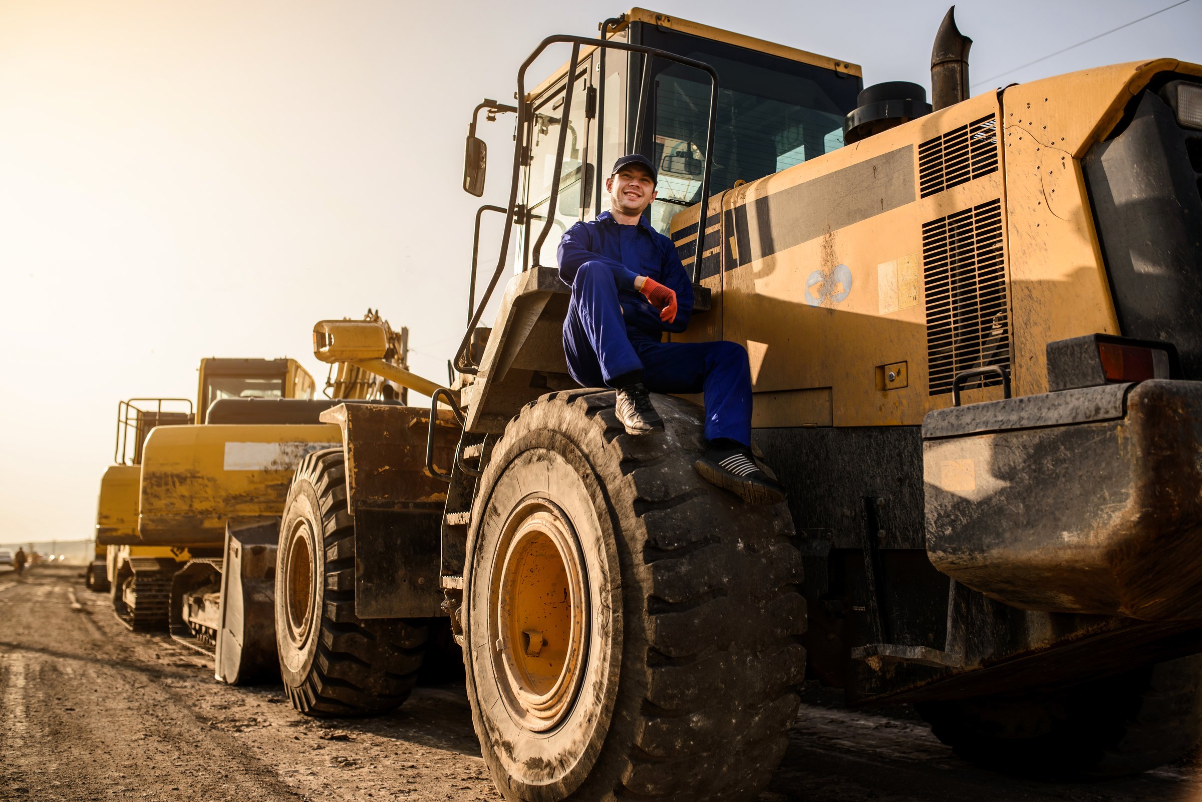 In the construction site a young man contractor builder in blue overalls is sitting on the bulldozer wheel and having a rest on sunset. Construction machinery concept