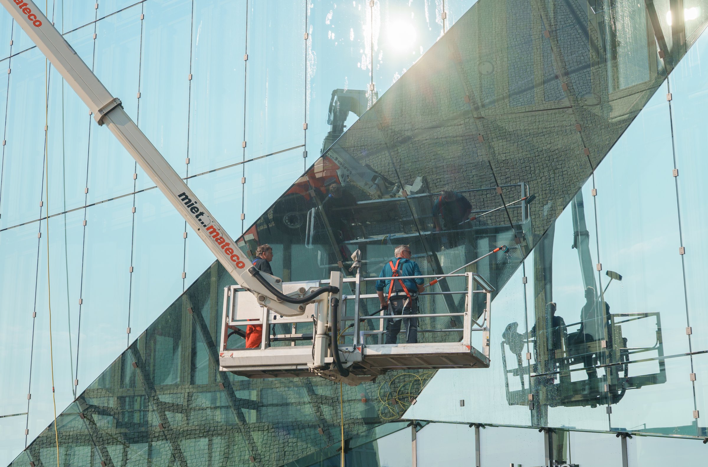 Berlin, Germany, 05.15.2025, Workers using a mechanical boom lift to clean or maintain the reflective glass facade of a modern building