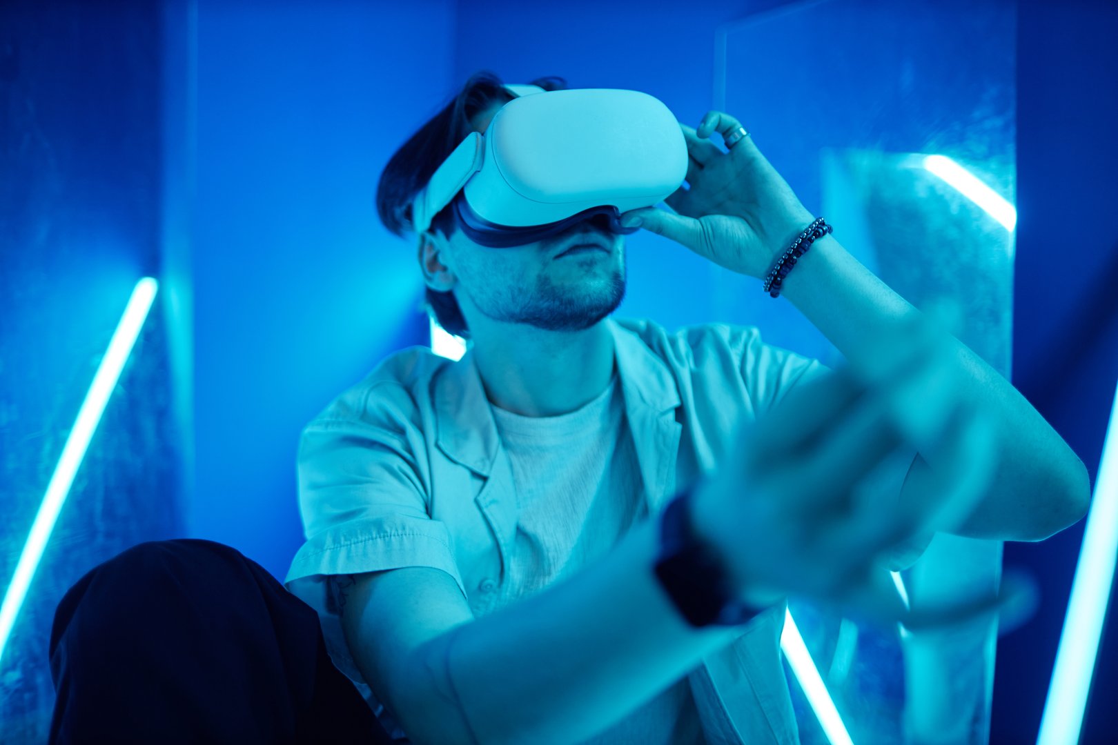 Portrait of man wearing VR headset sitting on floor in blue neon lights and reaching out experiencing digital cyberspace