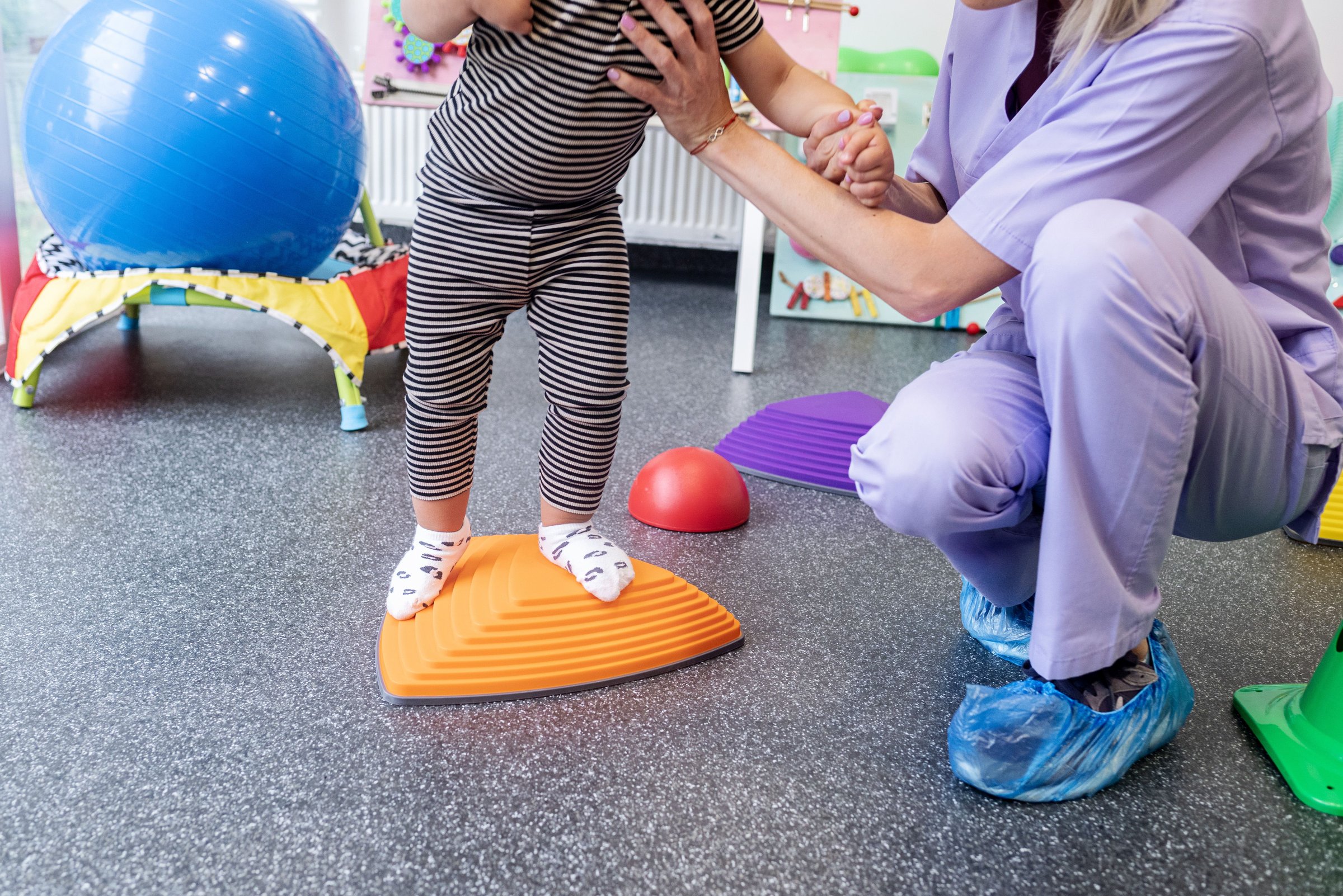 Child in striped outfit balancing on a sensory board with caregiver support in a therapy setting.