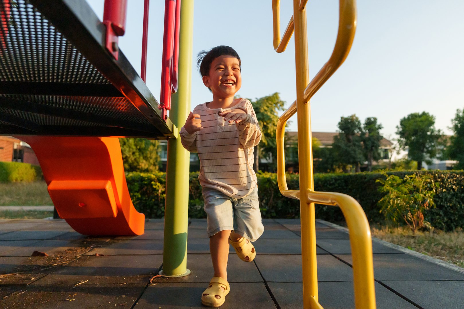 A young Asian boy runs excitedly through a colorful playground at sunset, enjoying the warm glow and vibrant surroundings. Laughter fills the air as he plays freely.
