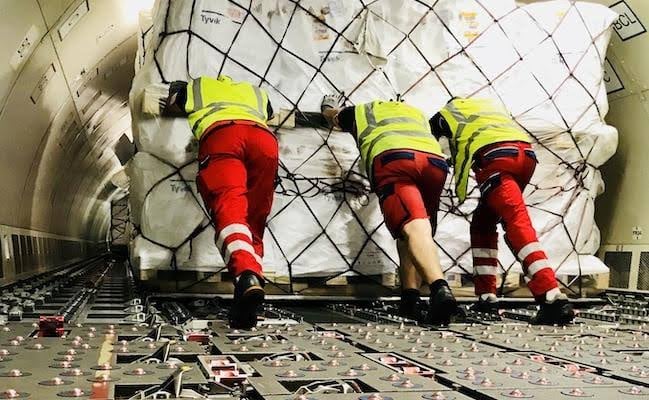 Three workers in safety vests and red pants push large cargo packages inside an airplane's cargo hold.