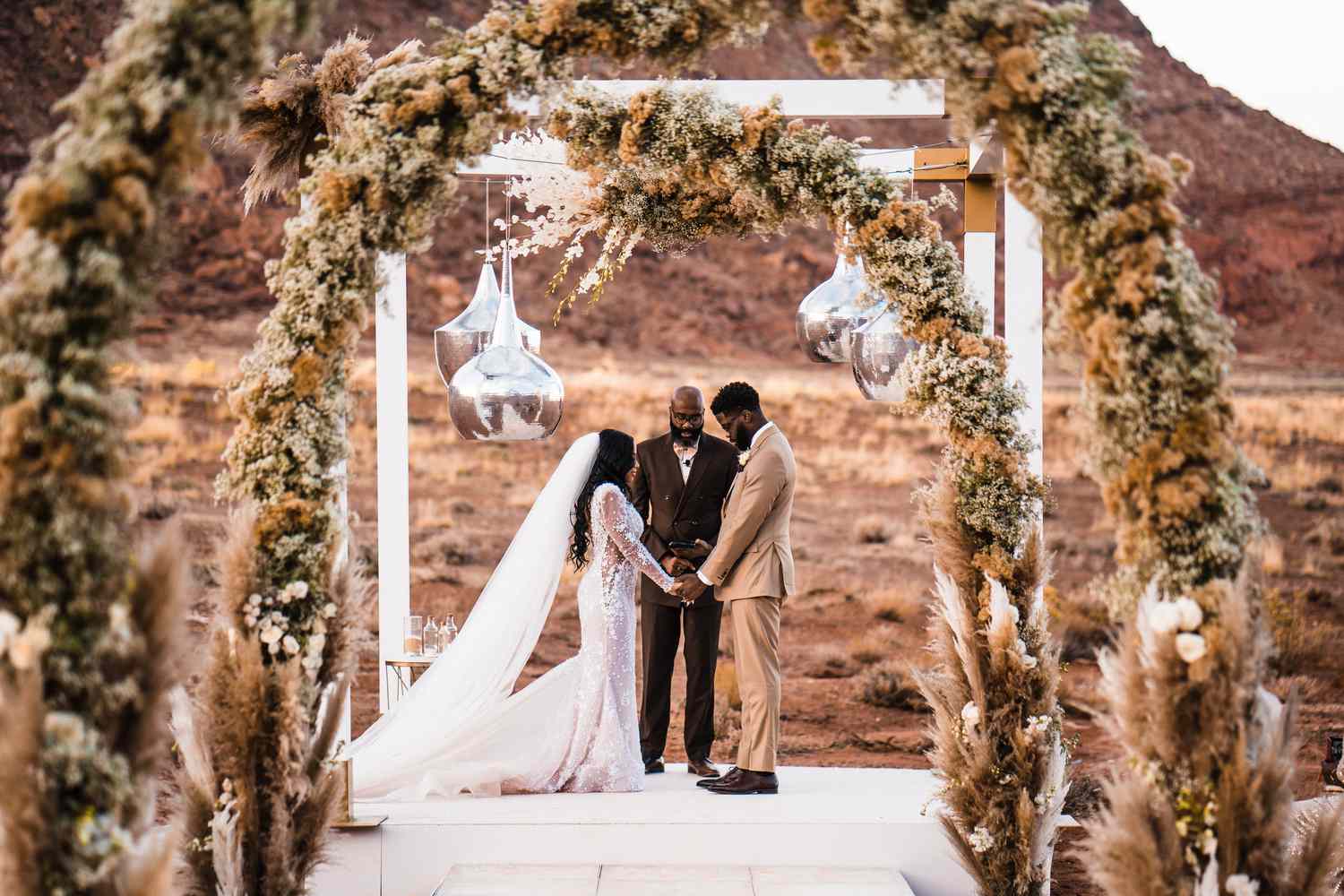 A couple exchanges vows in an outdoor desert wedding, under an arch adorned with pampas grass, officiated by a person in a suit.