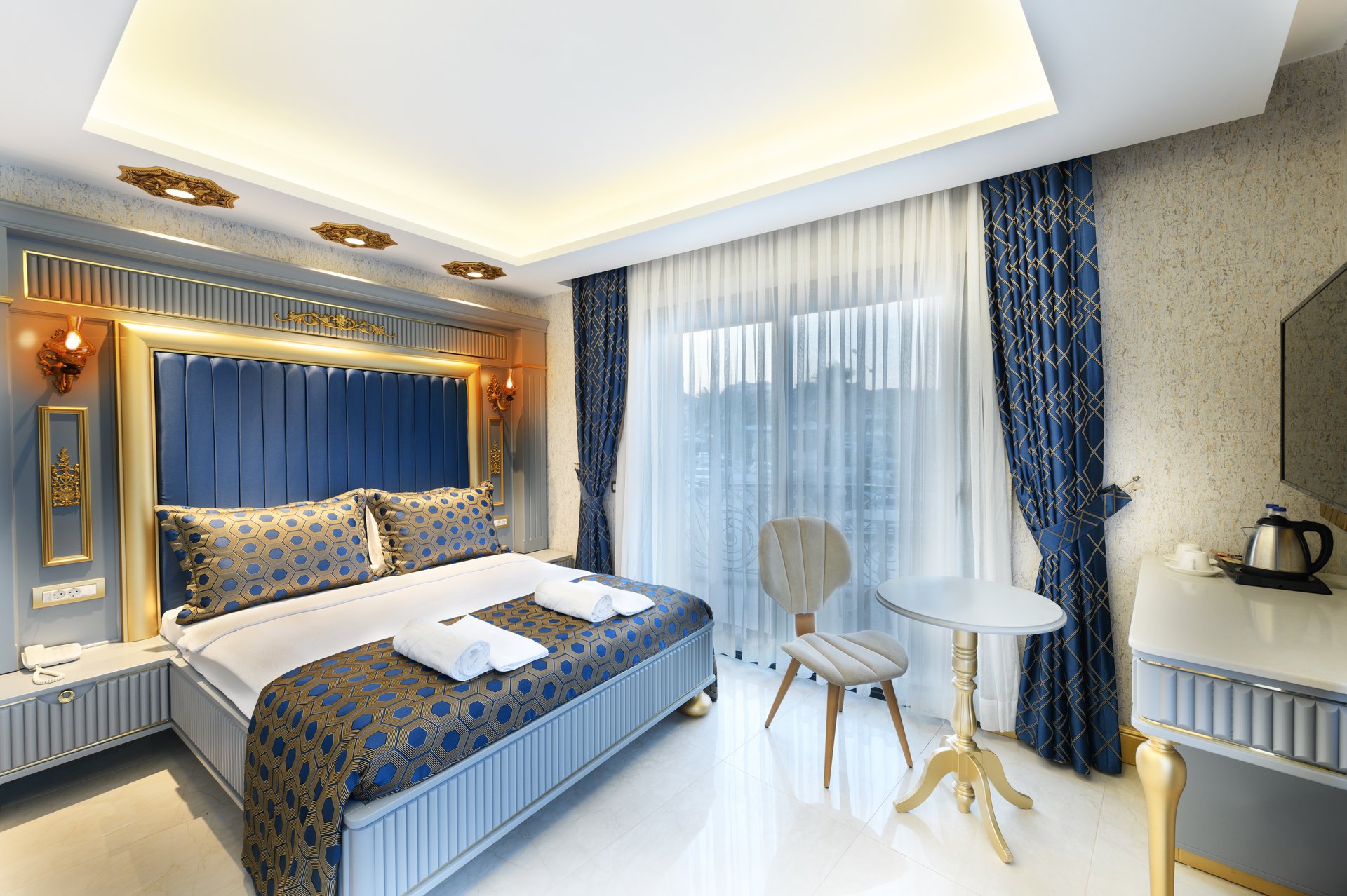 A photo of a single hotel room featuring a large bed, elegant decor, a wall-mounted TV, and a cozy seating area with natural light streaming through sheer curtains