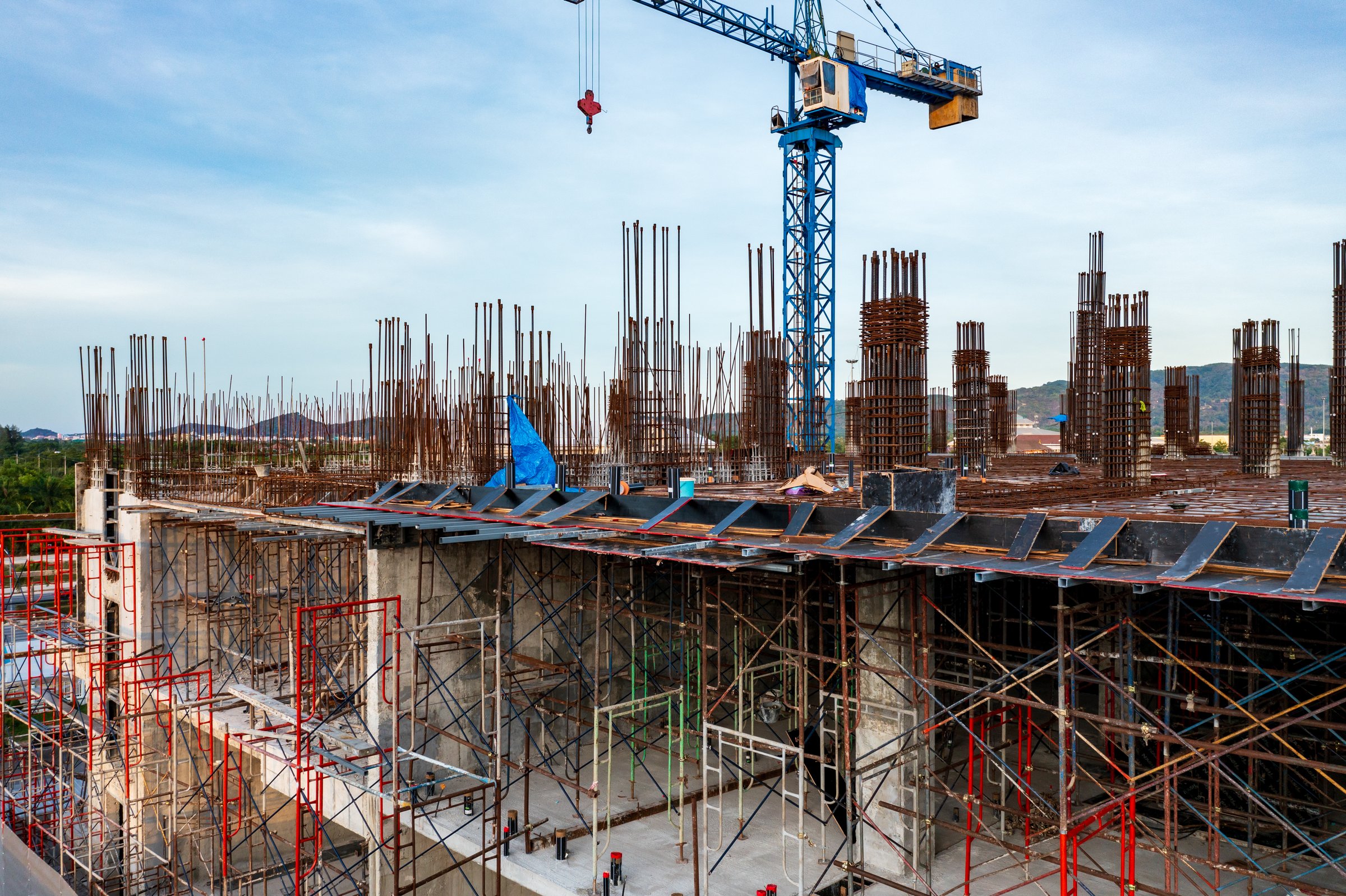 A construction site with a blue crane towering over reinforced concrete columns, steel rebar, and scaffolding, showcasing the progress of a building under development against a cloudy sky.