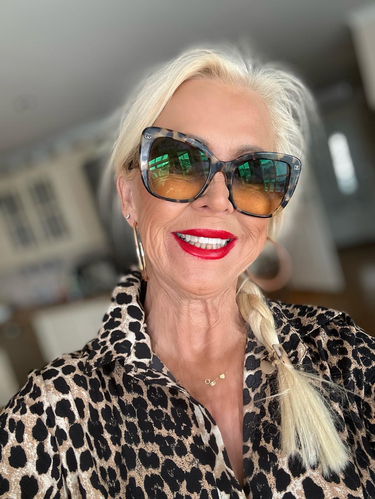 Woman with blonde hair wearing sunglasses and a leopard print outfit, smiling. Red lipstick and braid styled hair.