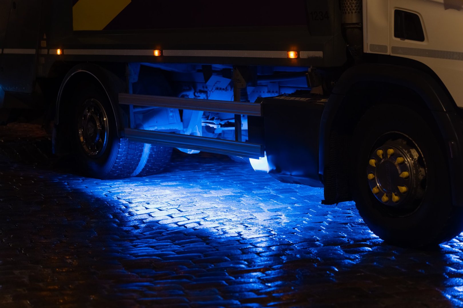 Blue underglow lighting shining beneath large vehicle on reflective cobblestone pavement highlighting wheels, suspension and lower frame