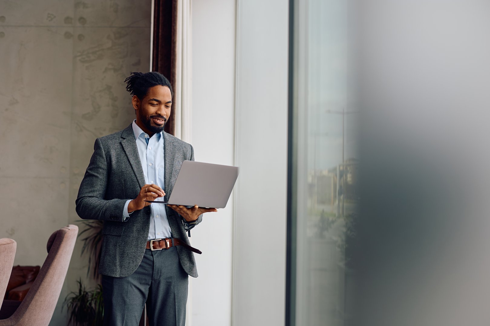 Happy African American businessman using laptop while standing by the window at his office. Copy space.