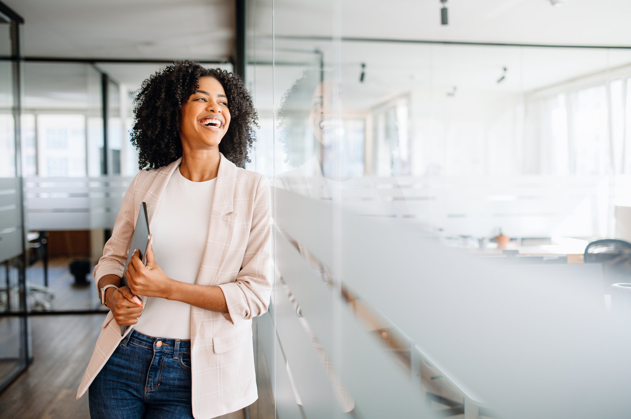 Brazilian businesswoman with an infectious smile standing in a modern office corridor and holding her laptop. Cheerful female employee satisfied with her occupation, feels happy and successful