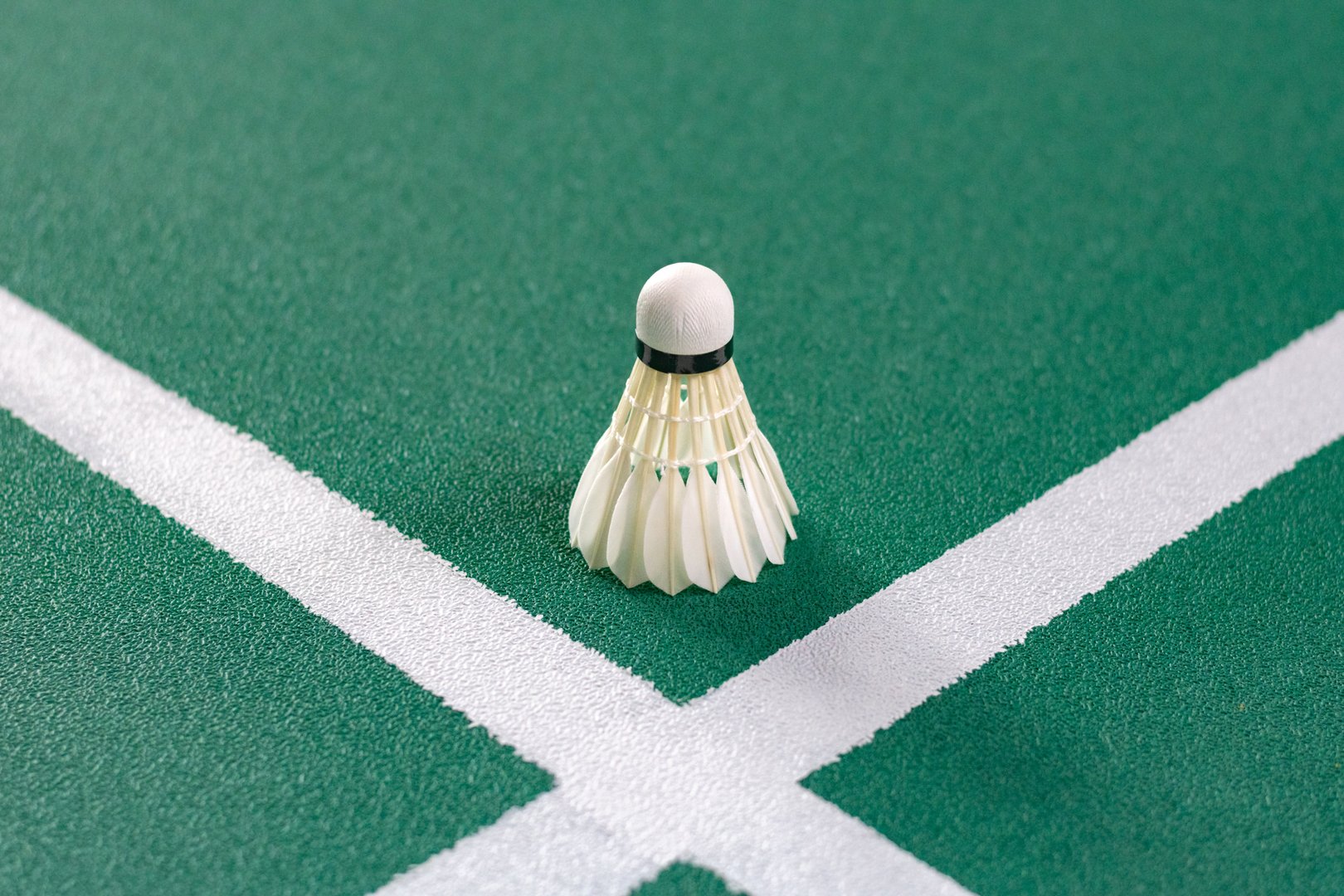 A perfectly positioned shuttlecock rests on the green court where two white lines intersect, capturing a peaceful moment of stillness before the next powerful serve in a badminton game.