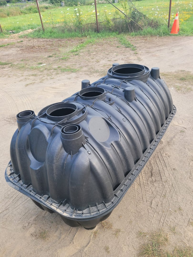 Large black plastic tank with multiple openings, sitting on sandy ground with grass and a traffic cone in the background.