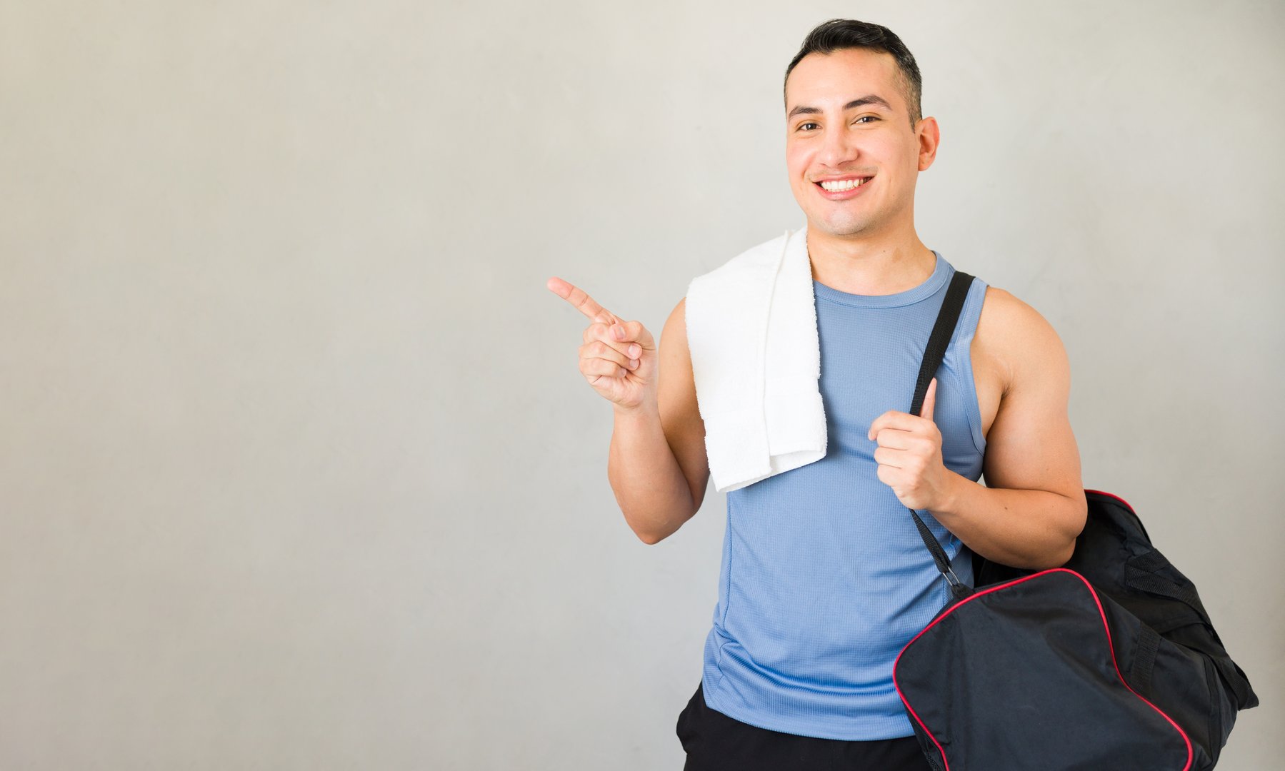 Athletic man wearing sportswear is holding a gym bag and pointing with a finger to the side while smiling