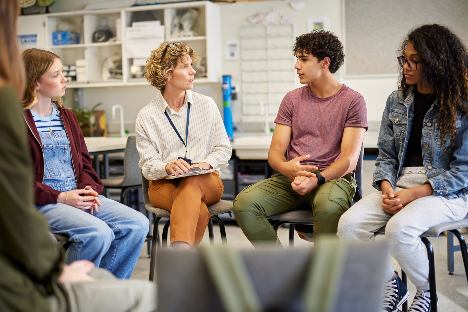 Multiethnic teenagers sitting in a circle engaged in an open group conversation to psychological support person. Instructor leading a thoughtful discussion with students in a relaxed setting. Mature woman supporting a group of girls and guys during a group therapy meeting session, giving advice.