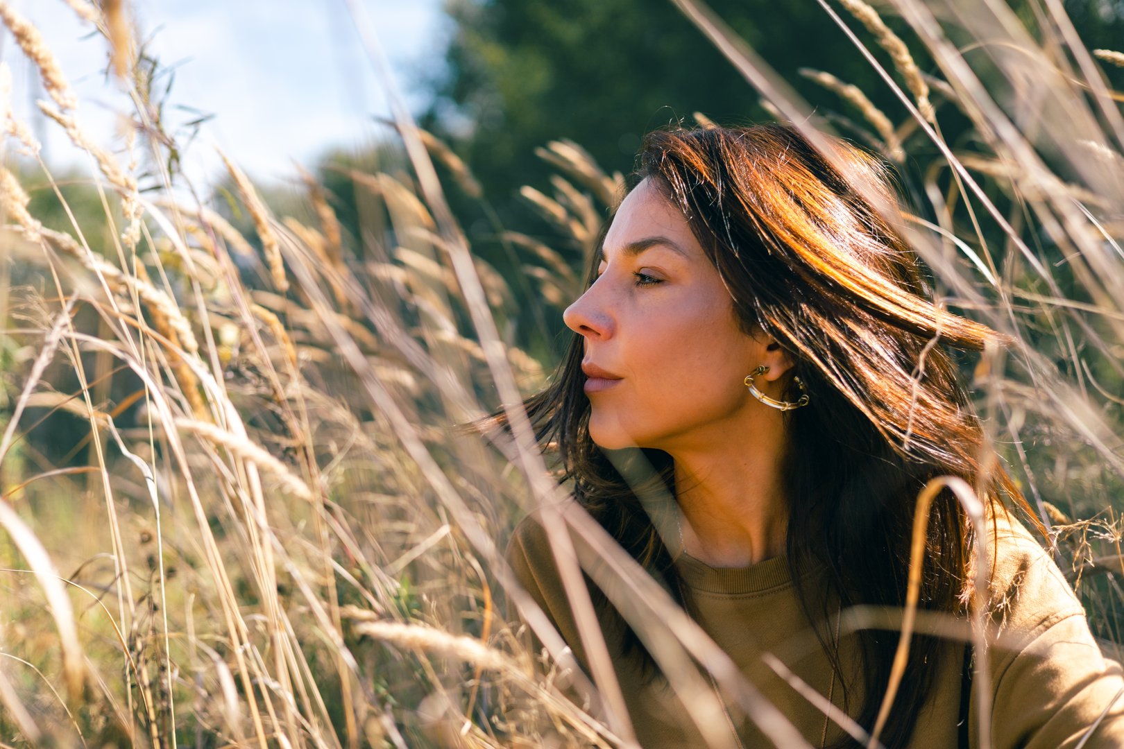 Side view of woman sitting among tall dry grass on a sunny autumn day. Concept of harmony, mindfulness, beauty, freedom, and connection with nature in soft warm natural light and calm mood.