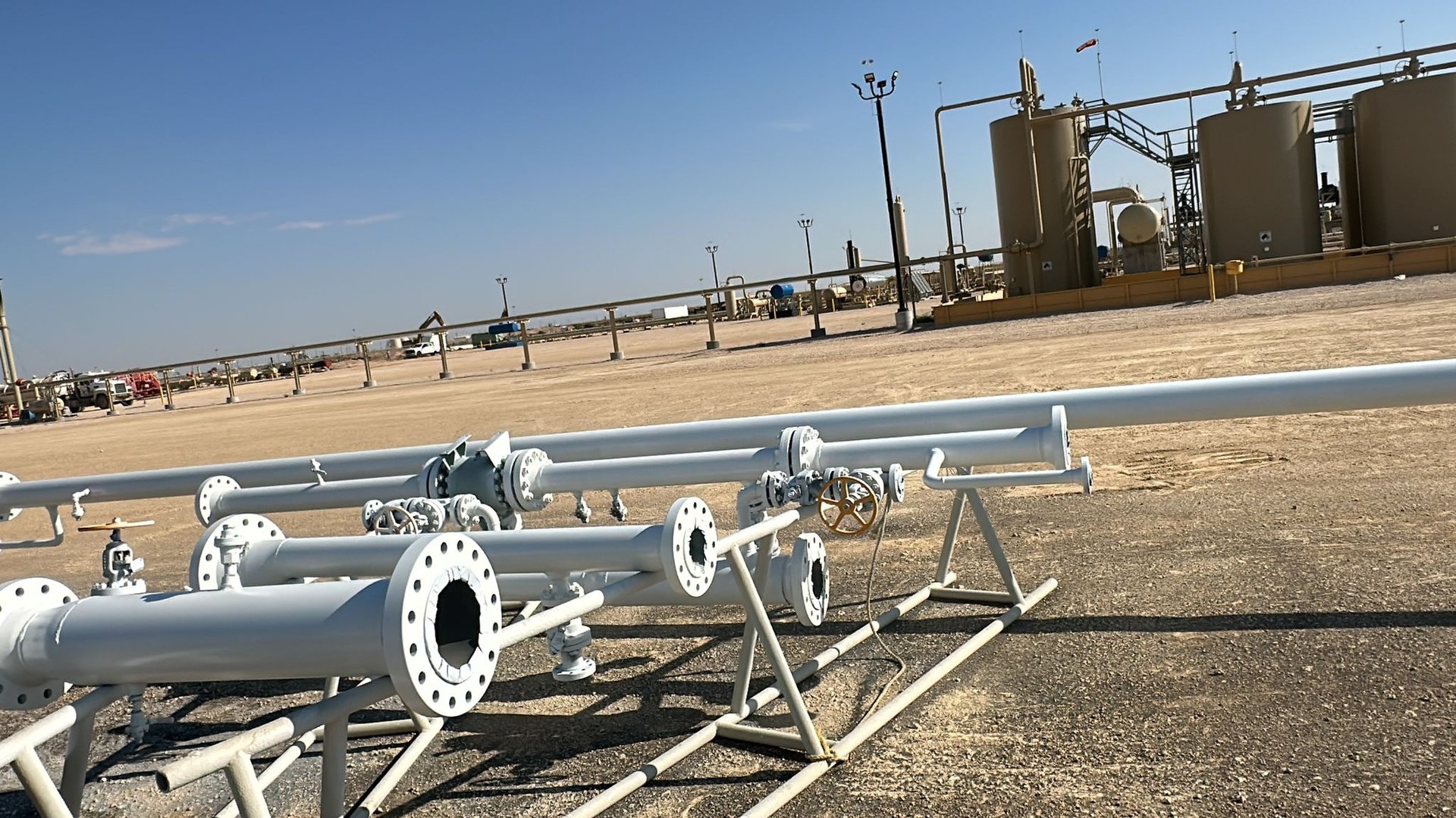 Industrial landscape with white pipe structures in the foreground and storage tanks in the background under a clear sky.