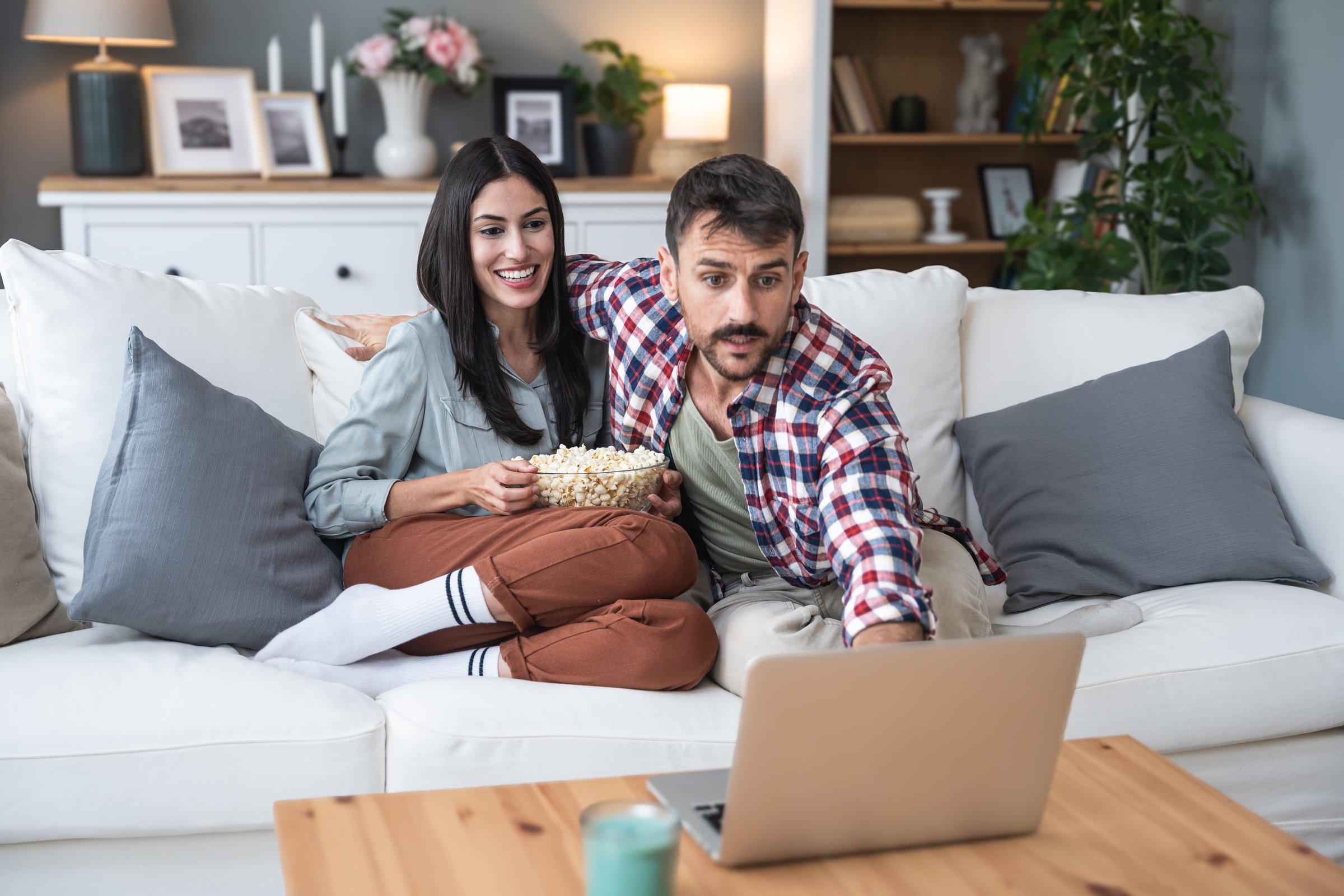 Young happy couple, simple living, sitting at home on sofa, choosing and watching movie on laptop computer, eating popcorn and enjoy time together. Healthy relationship lifestyle concept.