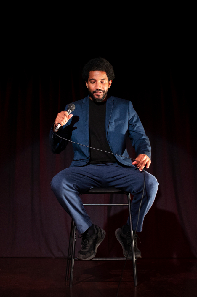 Man in blue suit sits on a stool on stage, holding a microphone, against a dark curtain backdrop.