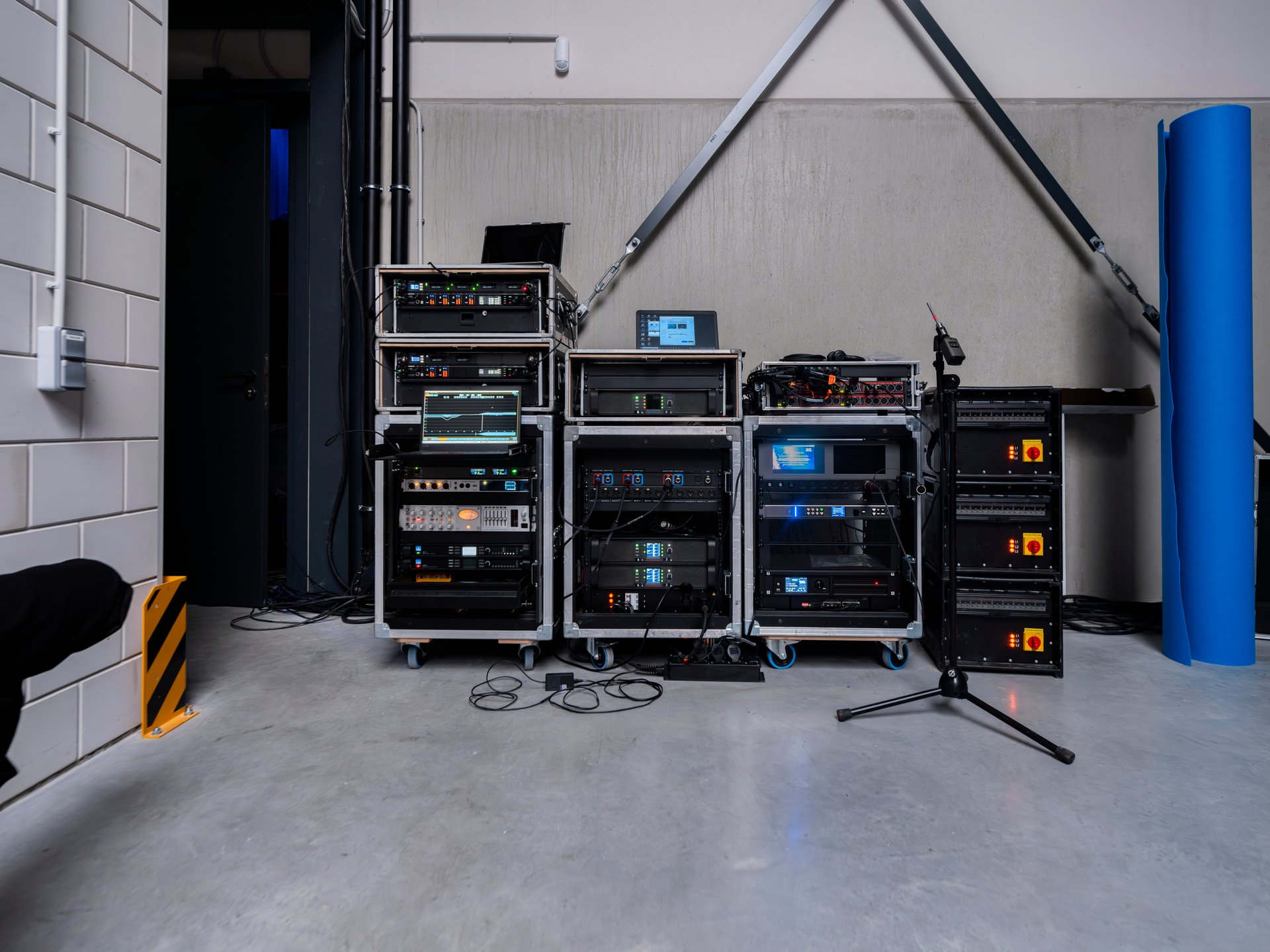 Mobile equipment rack filled with professional audio gear including amplifiers, processors, and control units with LED displays and status lights. Black aluminum stage truss structure positioned above the rack system in modern production facility.