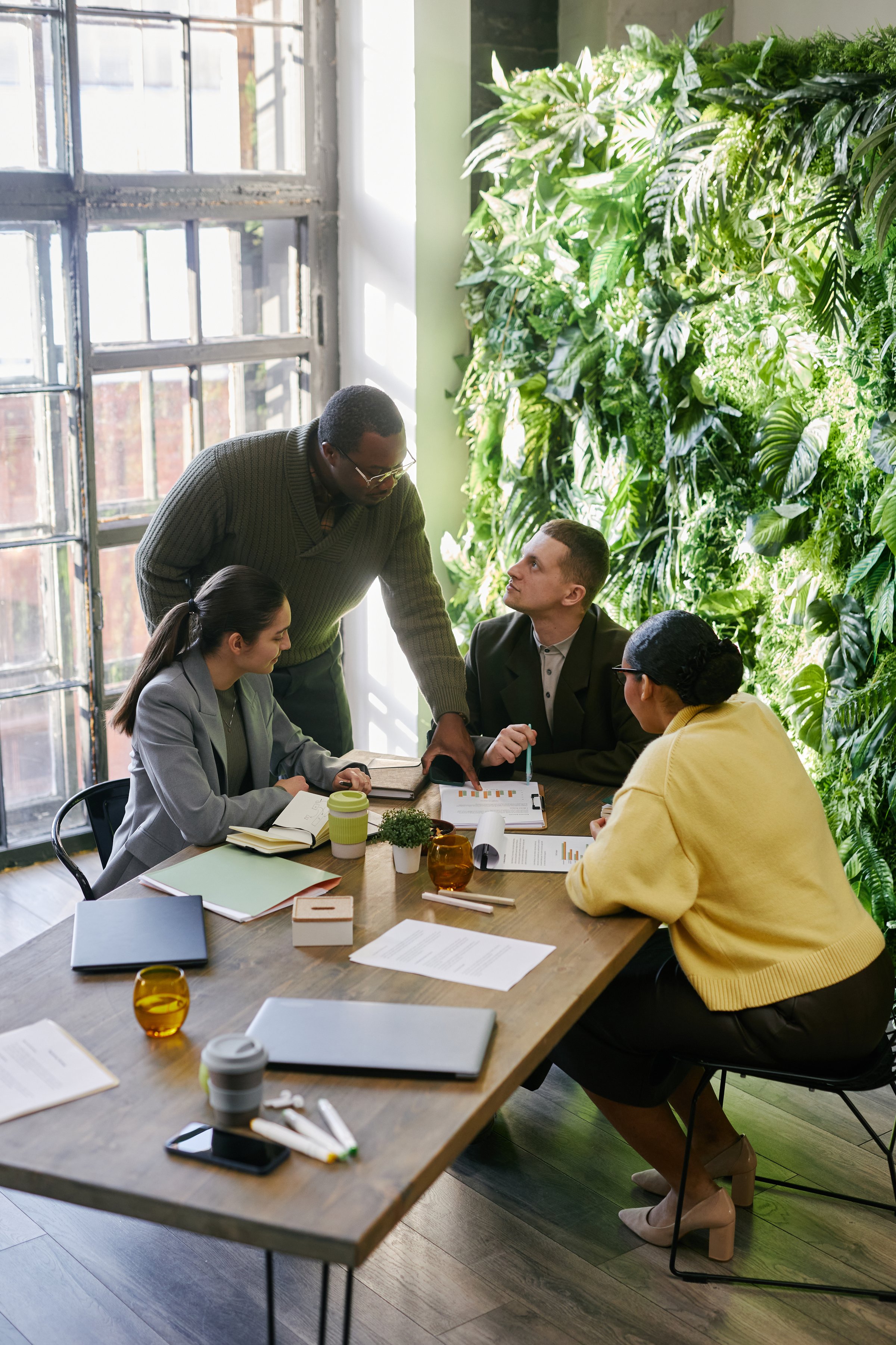 Vertical shot of diverse group of young adult professionals collaborating around table in modern office, Black man standing and explaining business strategy to colleagues during meeting