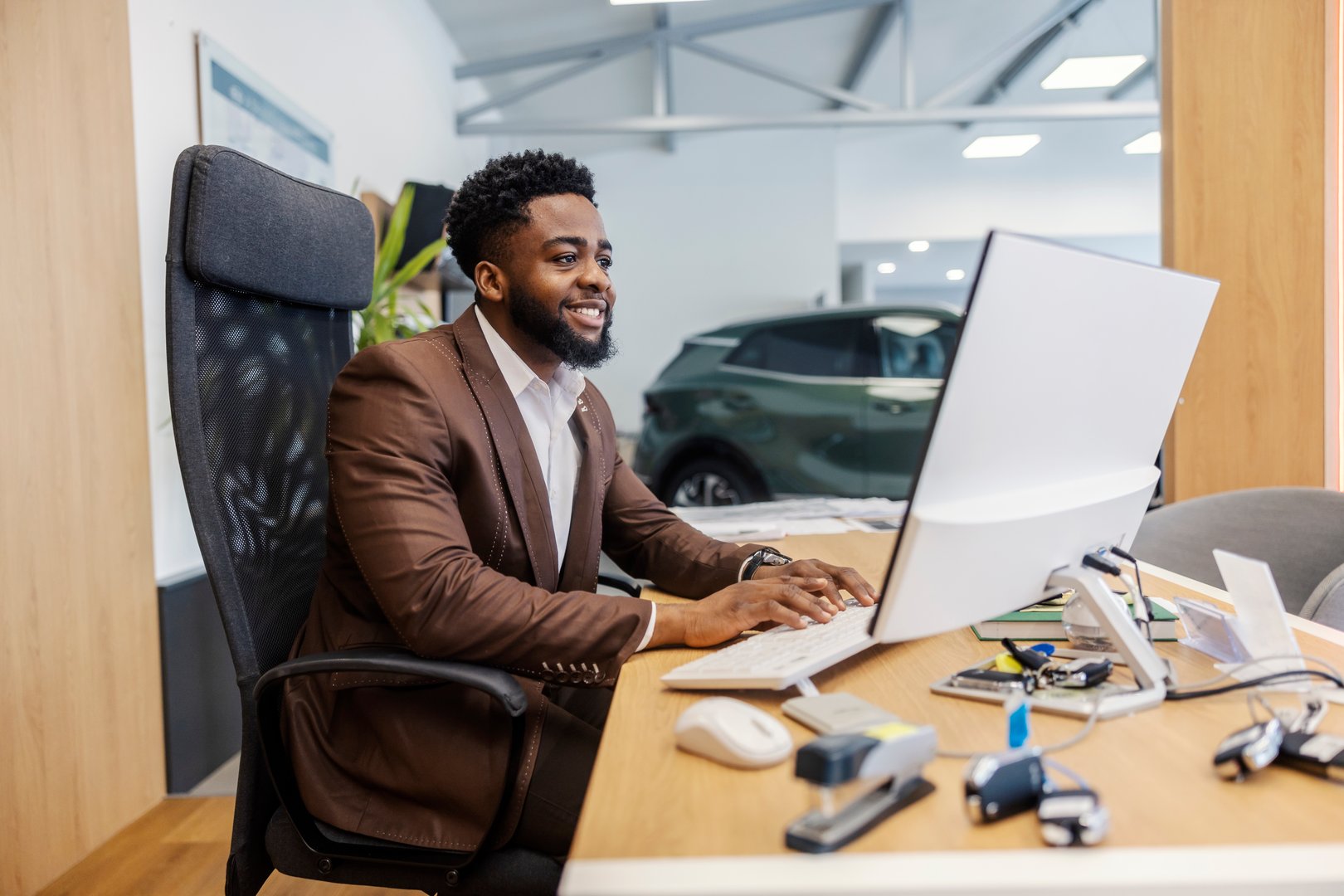 Multicultural car dealer sitting at car showroom and typing contract on computer keyboard while smiling at computer.