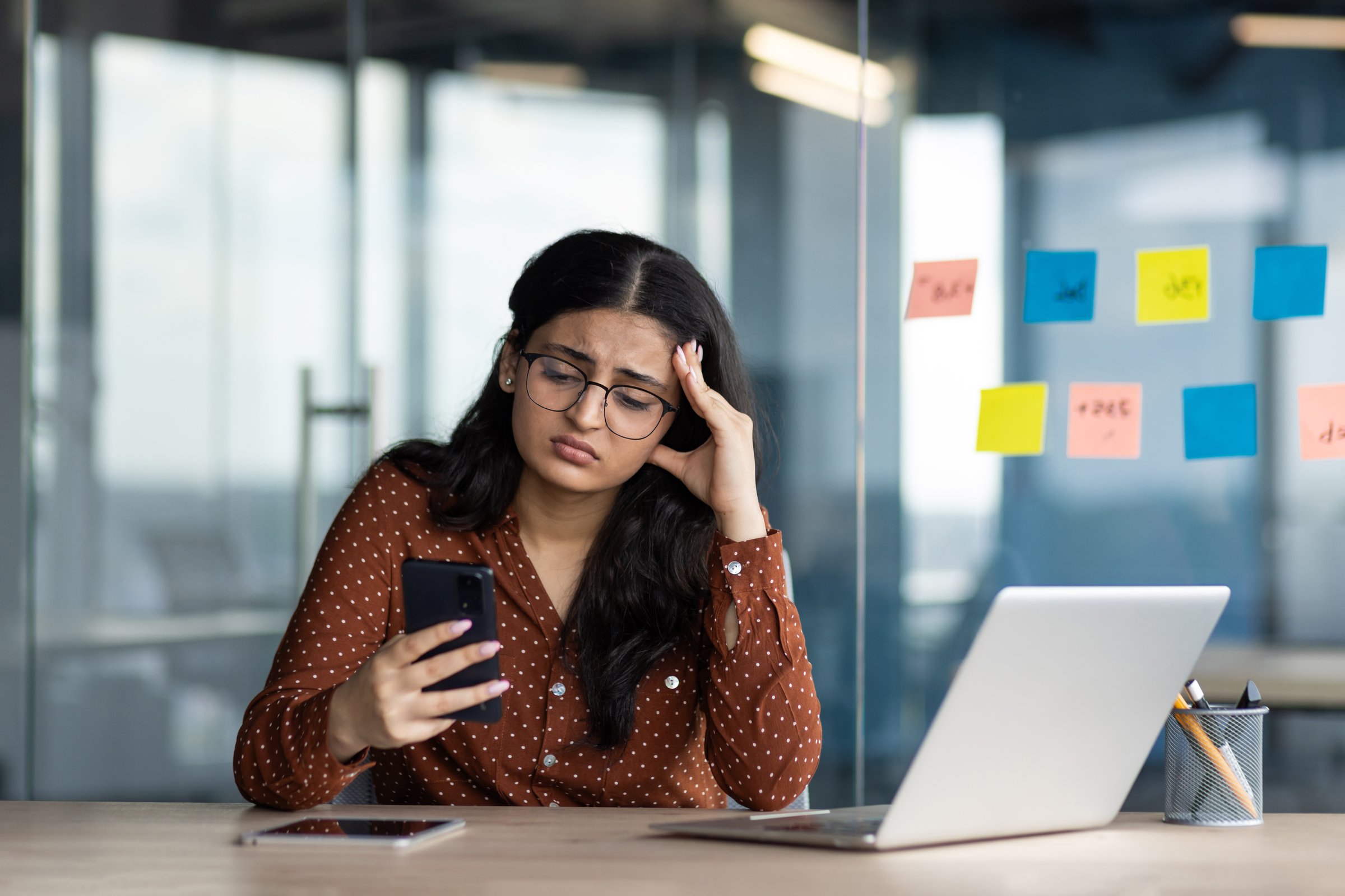 Sad disappointed business woman holding phone in hands, office worker received message with bad news online, latin american woman unhappy with business result inside building.