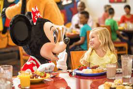 A child dining with Minnie Mouse at a restaurant, smiling and enjoying a plate of food. Others are visible in the background.