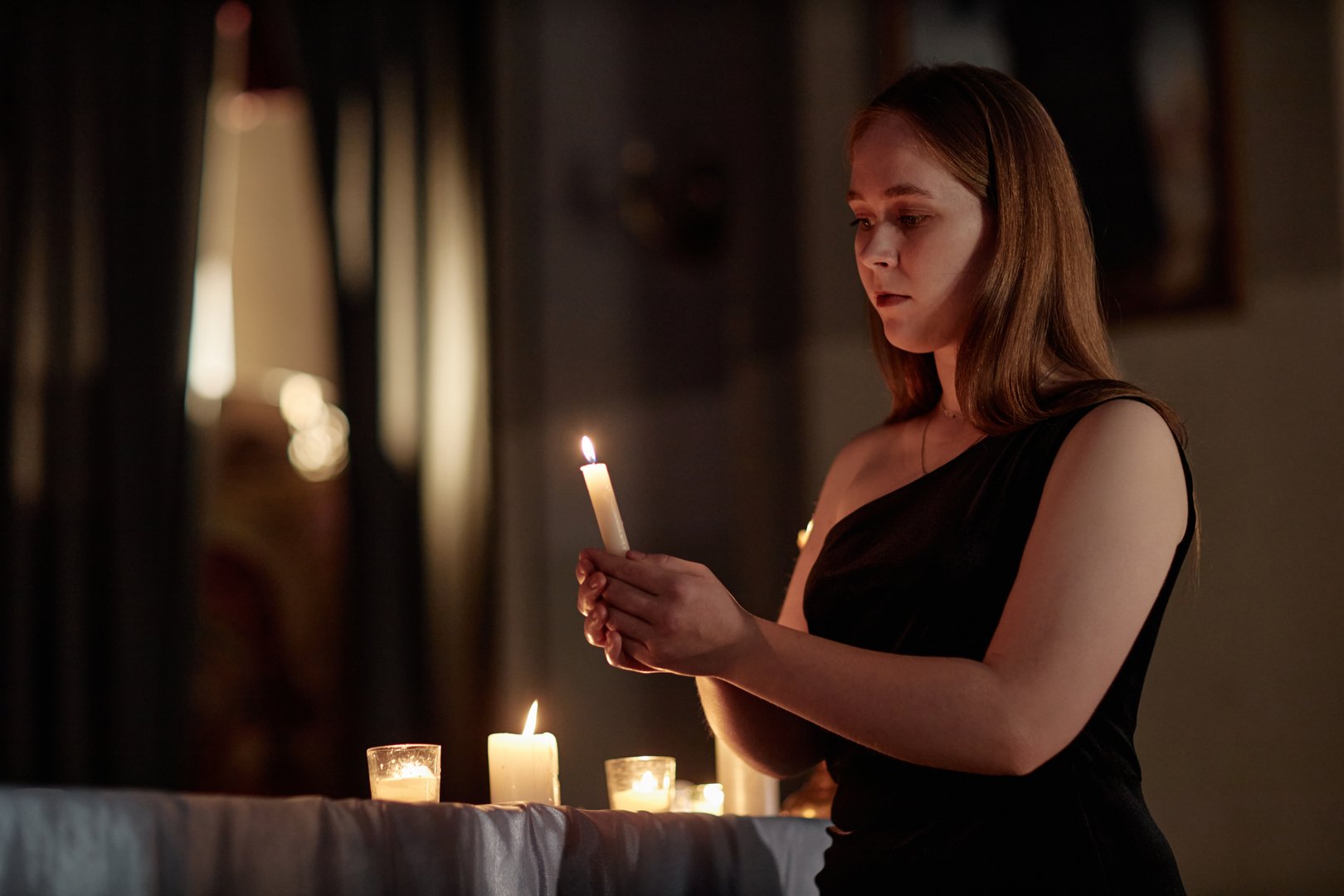 Medium shot of young woman in elegant black dress putting out candle while praying in church, copy space