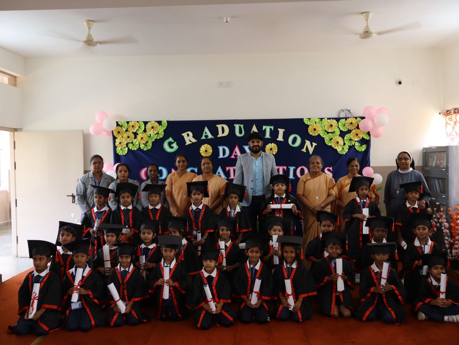 Children in graduation caps and gowns pose with adults in front of a "Graduation Day" banner in a decorated room.