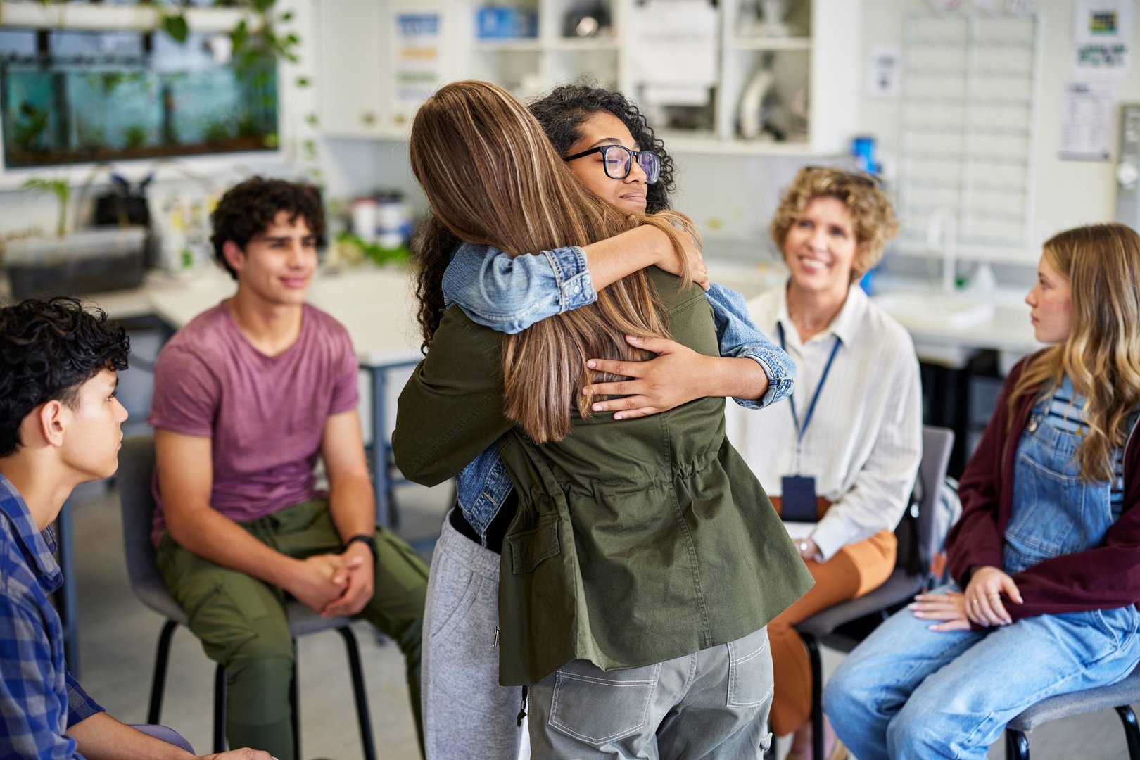 Two female students embracing during a group counseling session at high school, offering comfort and emotional connection. Multiethnic classmates observe moment of support with attentive and compassionate expressions. Therapist smiling in the background, witnessing positive outcome of the session.
