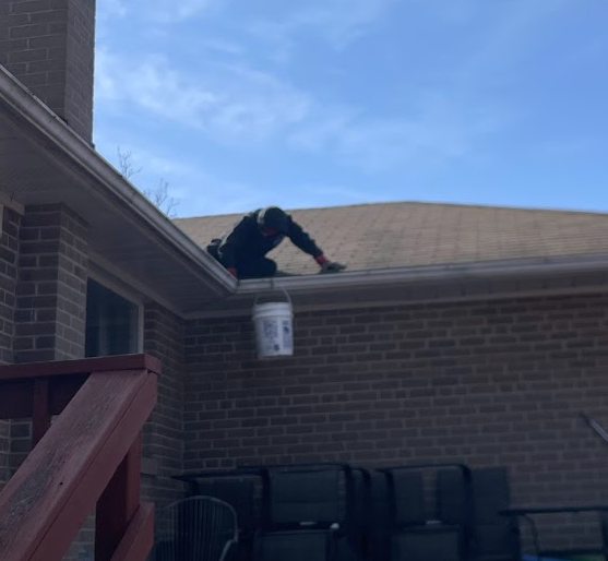 Person on a roof reaching for a hanging bucket, with outdoor furniture visible below on a brick house porch.