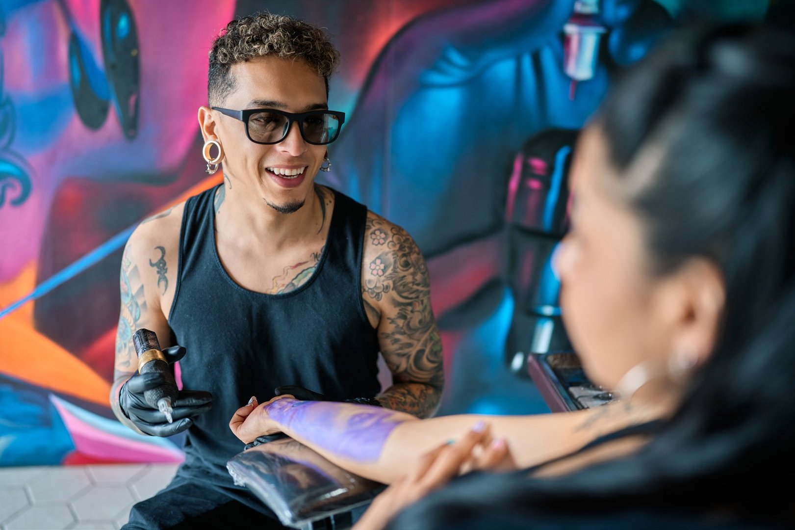 Professional tattoo artist smiling while skillfully tattooing a woman's arm in a vibrant, colorful studio filled with creative energy
