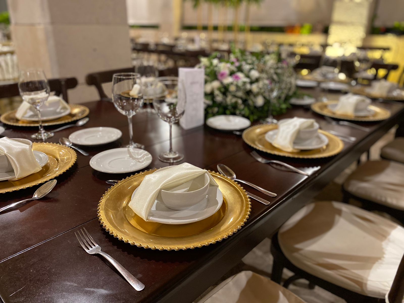Elegant table setting with gold chargers, white plates, and wine glasses on a dark wooden table. Floral centerpiece visible.