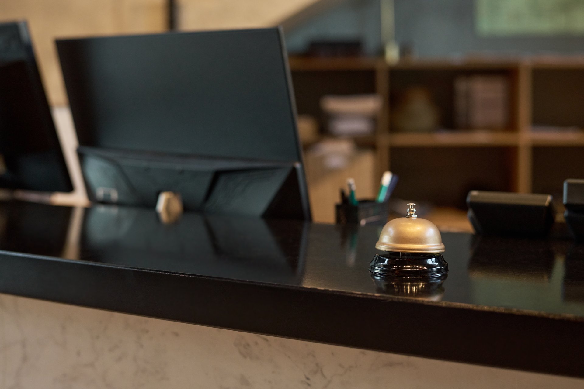 Modern hotel reception desk featuring a vintage silver bell and sleek black computer monitors. Background includes shelves with books and decorative items creating welcoming atmosphere