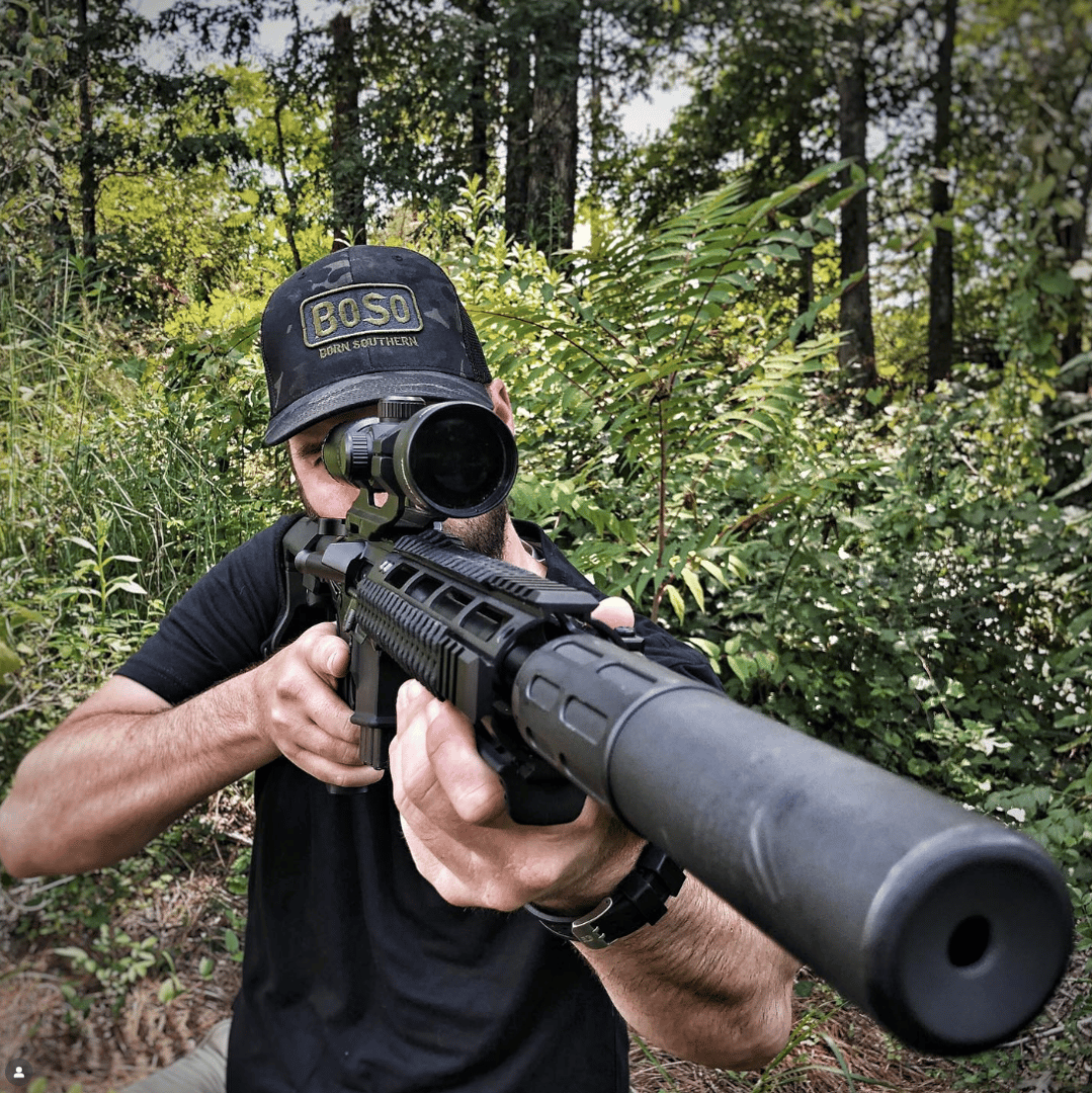 Person aiming a rifle with a scope in a wooded area, wearing a black cap and shirt, surrounded by green foliage.